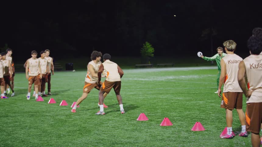 A team of young male soccer players practices one-on-one defensive agility drills on a lit field at night