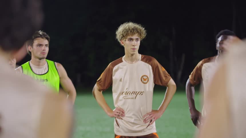 A diverse group of young soccer players attentively listens to their coach during a team huddle on the field at night