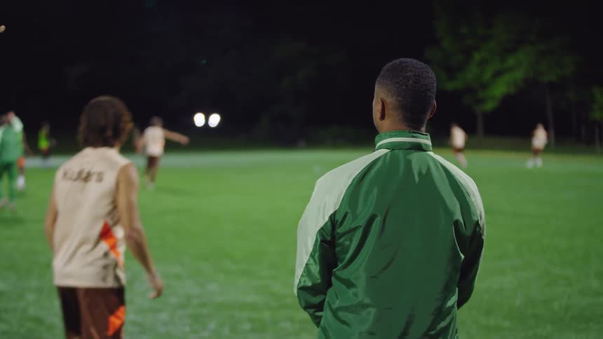 A soccer coach from behind wearing a green jacket watches his team practice on a field at night