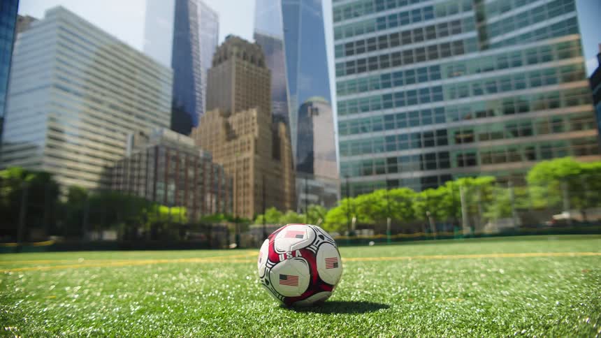 A patriotic soccer ball with the USA flag sits on a lush green sports field in a park surrounded by a modern city skyline