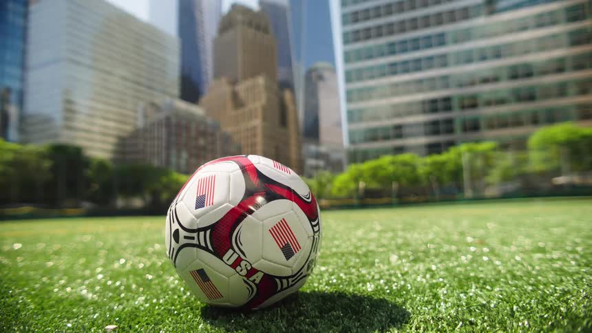 A camera tilts down from the iconic One World Trade Center and New York City skyline to reveal a USA soccer ball on a green field