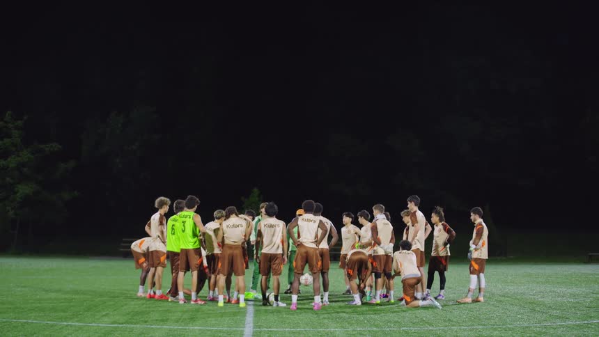 A male soccer team huddles together on a brightly lit field at night for a strategic team meeting with their coach