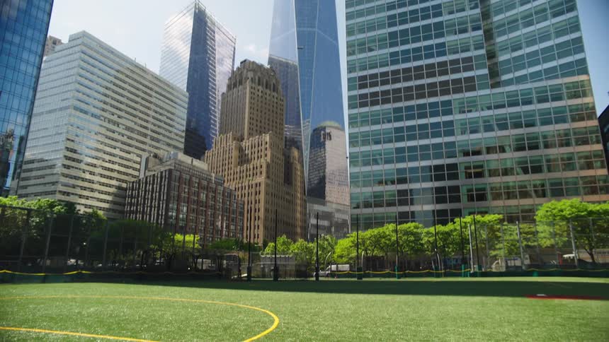Tilting down from sunlit skyscrapers in New York City's Financial District to reveal an empty green sports field