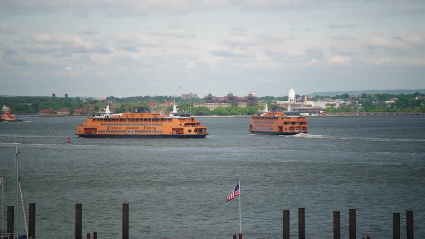 Two iconic Staten Island ferries travel across the New York Harbor on a cloudy day with an American flag in the foreground