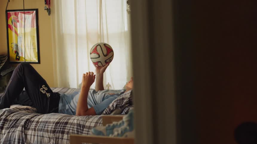 A reveal shot shows a young man relaxing on his bed in his sunlit bedroom, spinning a soccer ball