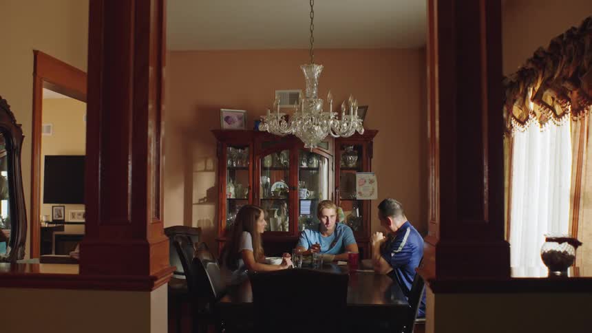 A family sits together at a formal dining room table for a serious conversation in a dimly lit room