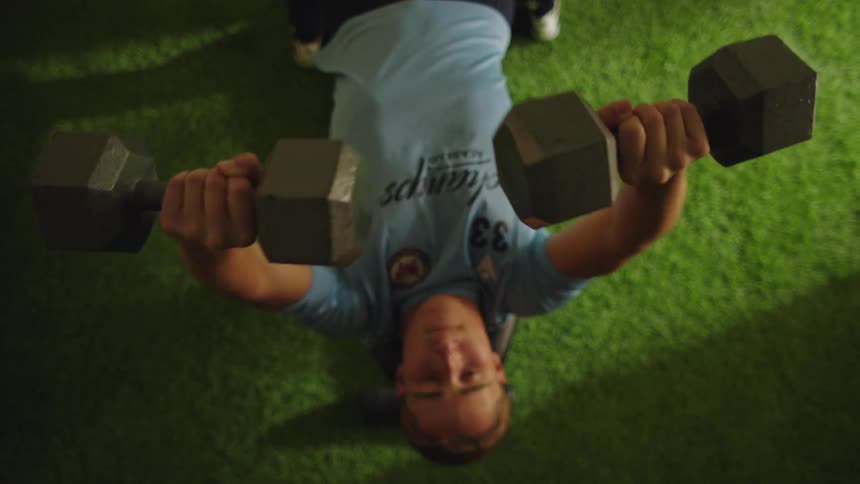A male athlete from a top-down perspective does a dumbbell chest press while lying on a weight bench over artificial turf