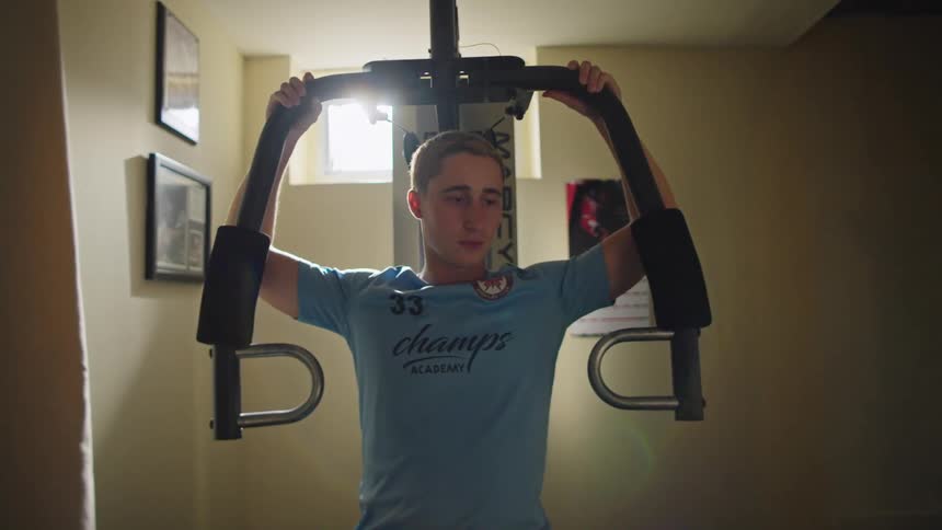 A focused young man performs chest exercises on a weight machine in his home gym, with a cinematic lens flare from a window in the background