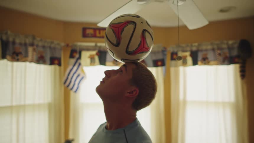 A talented young man skillfully balances a soccer ball on his head in slow motion while practicing in his bedroom