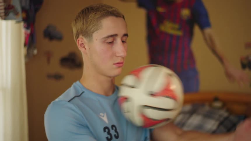 A contemplative teenage soccer player in his jersey sits on his bed, tossing a soccer ball between his hands in slow motion