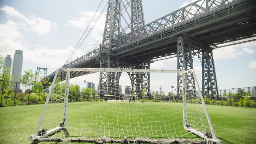A slow tilt down reveals a soccer field and goal located directly beneath the Williamsburg Bridge in a sunny urban park
