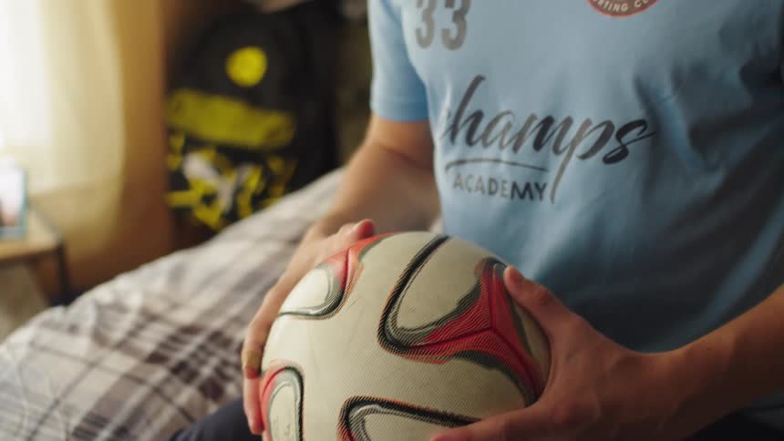 A young soccer player sits on his bed, holding a soccer ball with a focused expression on his face