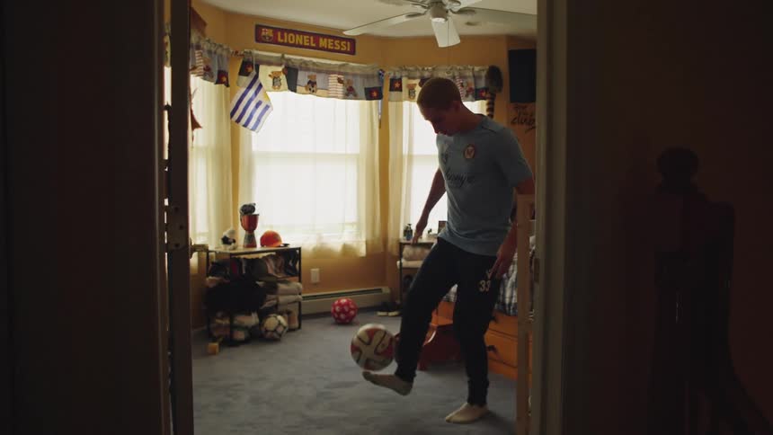 A dedicated young soccer player practices juggling a ball in slow motion inside his bedroom filled with sports memorabilia