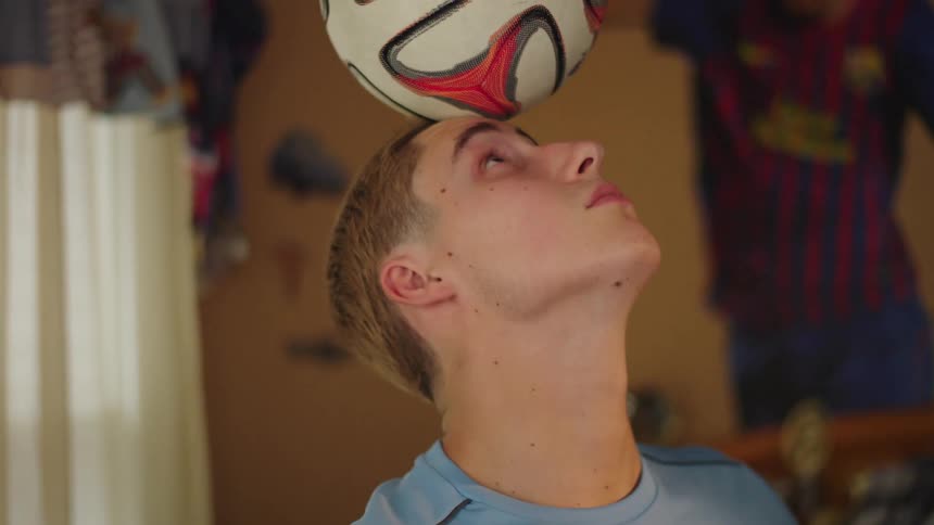 An aspiring young athlete hones his soccer skills, intensely focused on balancing a ball on his head in his bedroom