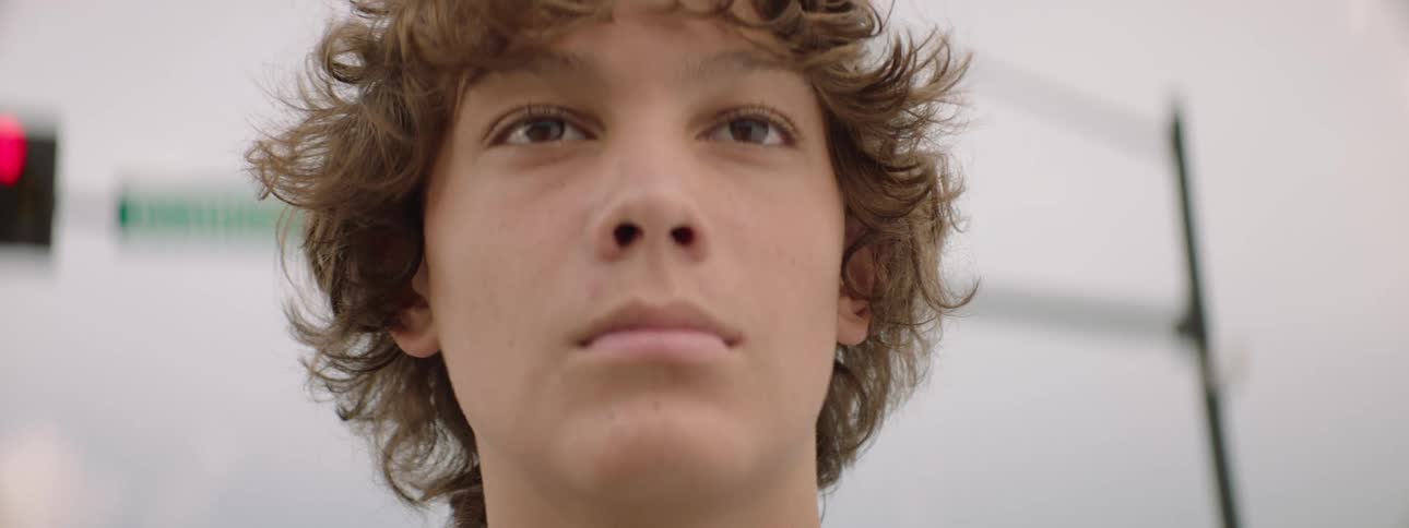 A focused teenage boy with curly brown hair looks directly at the camera in an intense close-up shot