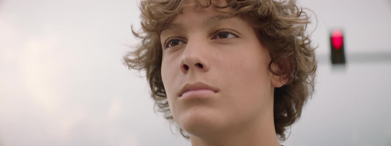 A thoughtful teenage boy with curly brown hair looks off into the distance in a slow-motion close-up