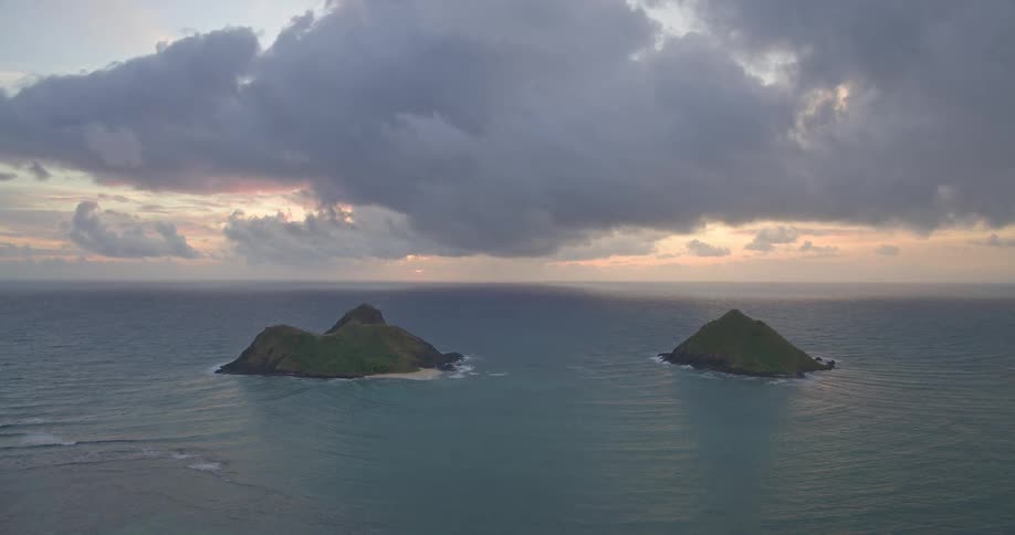 The iconic Mokulua Islands sit peacefully in the Pacific Ocean under a dramatic, cloudy sunrise sky in Hawaii