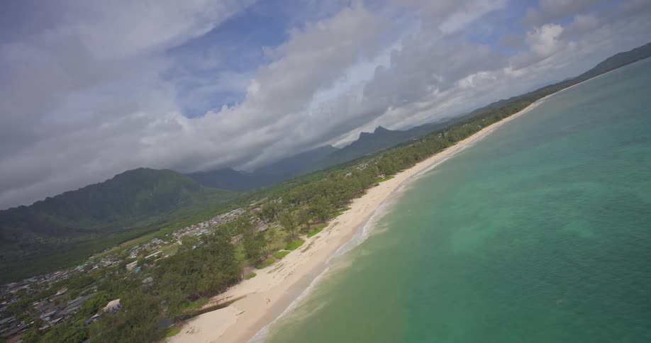 A breathtaking aerial descent over a pristine tropical beach reveals turquoise waters meeting the sandy shore, with lush green mountains under a cloudy sky