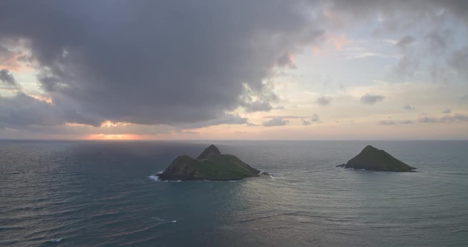 An aerial view pulls back from two lush green tropical islands in the ocean at sunrise under a dramatic cloudy sky