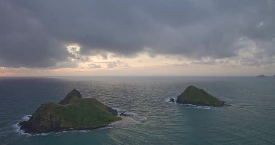 A dramatic wide aerial shot captures the iconic Mokulua Islands off the coast of Oahu, Hawaii, under a moody, overcast sky at sunrise