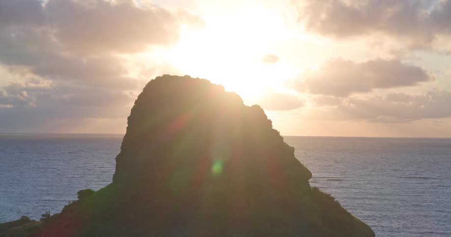 Aerial footage reveals a silhouetted tropical island as the sun rises, casting golden light and beautiful sunbeams across the ocean