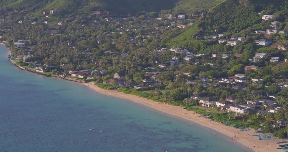 A slow aerial pan across a lush tropical coastline in Hawaii, revealing a beautiful beach, clear blue ocean, and homes built into the green mountainside