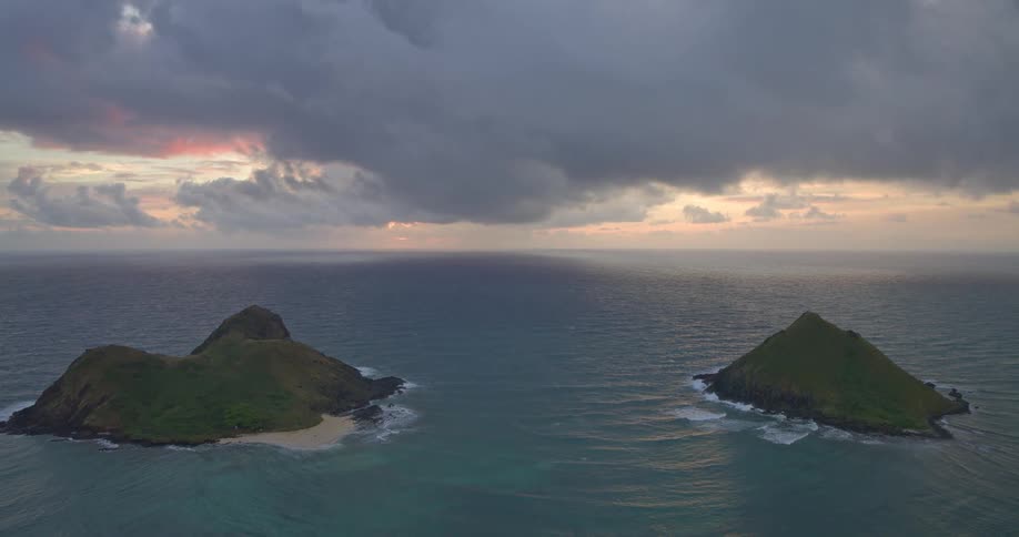 A dramatic, cloudy sunrise casts a soft glow over two lush green tropical islands in the vast ocean