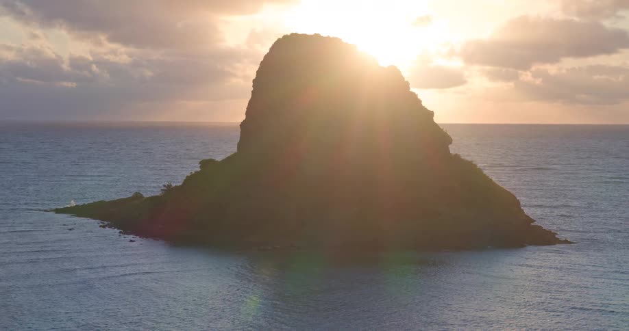 The sun's bright rays emerge from behind a silhouetted tropical island as it rises over the tranquil ocean