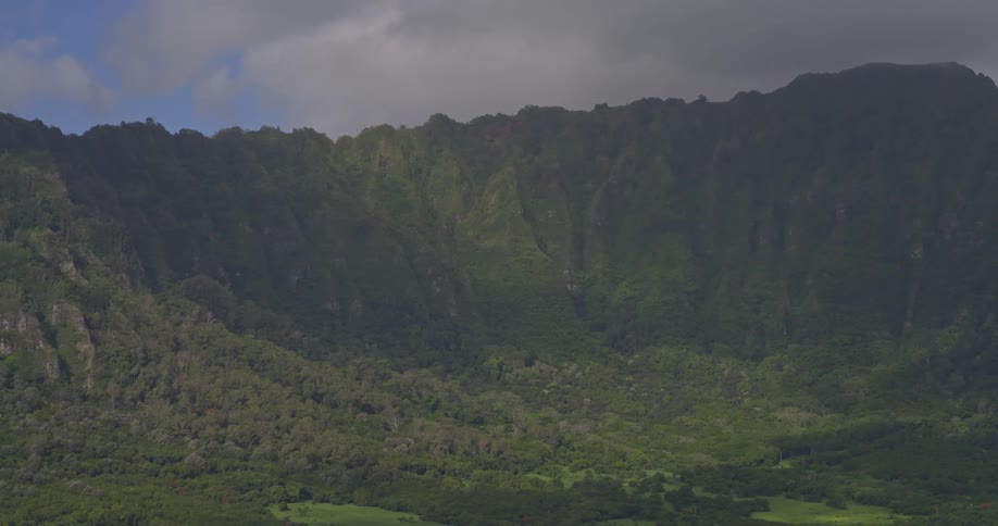 A slow zoom out of a majestic, lush green mountain range partially covered by the shadow of a large cloud