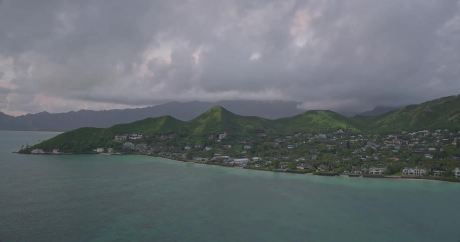 A serene aerial view of a Hawaiian coastal village situated between lush green mountains and the calm turquoise ocean under a moody, overcast sky
