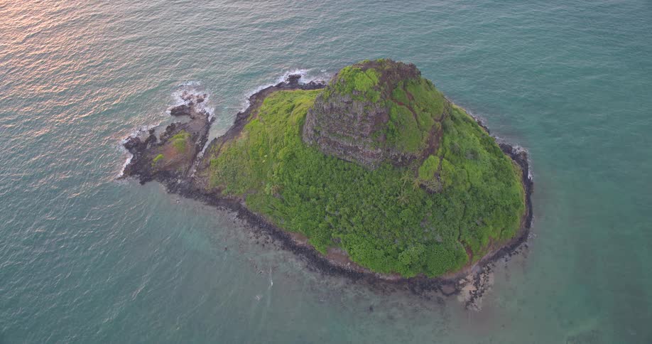 Slow aerial pull-back reveals the lush, green, and iconic Chinaman's Hat island off the coast of Oahu, Hawaii at sunrise