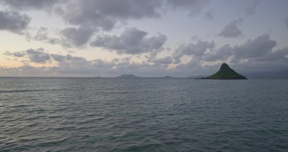 A tranquil seascape shows the gentle ocean lapping towards Mokoliʻi island under a soft, cloudy sky at dawn