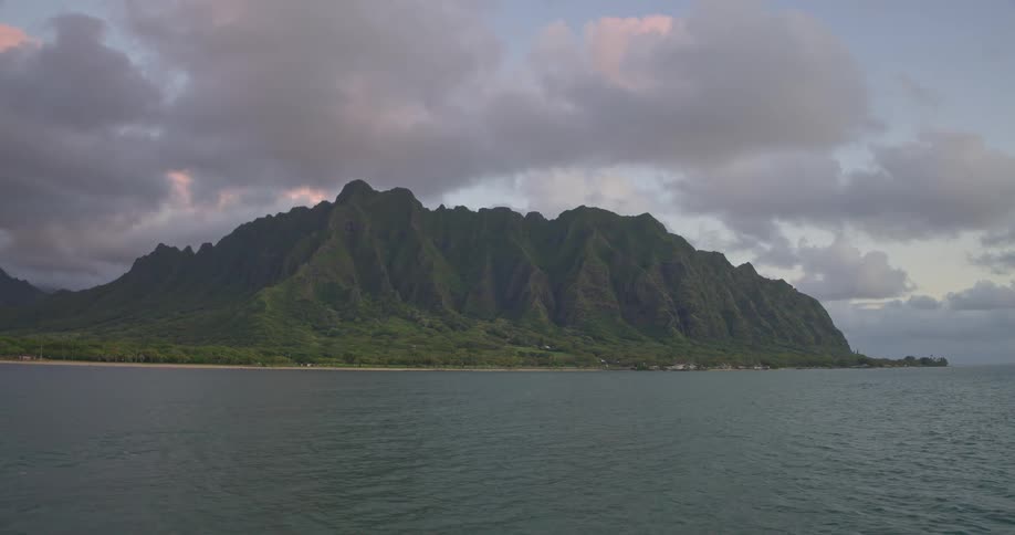 A serene wide shot of a lush green Hawaiian mountain range meeting the calm ocean under a cloudy sky at dusk