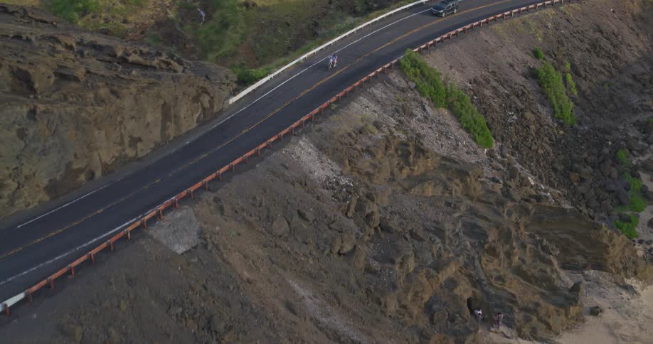 Aerial view follows a motorcycle with two riders as they cruise along a scenic coastal highway carved into a steep, rocky cliffside