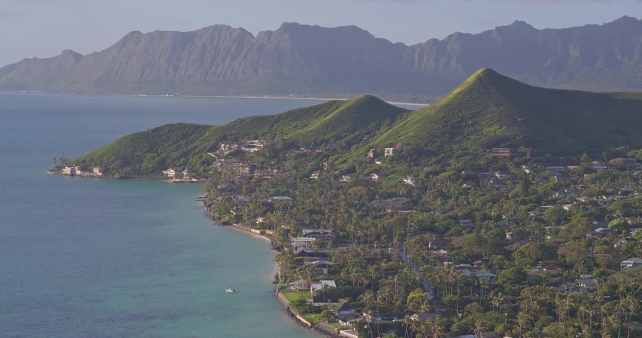 A slow zoom-out reveals the picturesque Hawaiian coastline, featuring a lush town nestled between green hills and a vast mountain range