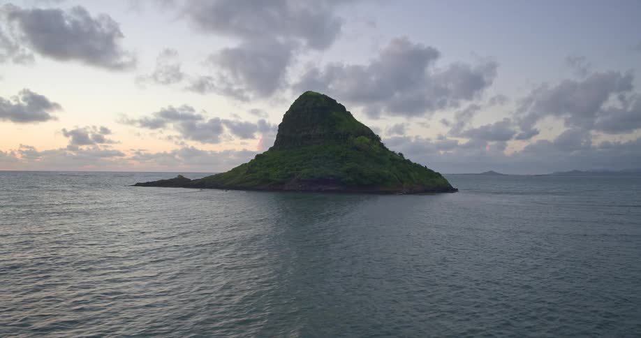 An aerial drone flies over the calm ocean towards a lush, green tropical island as the sun begins to rise