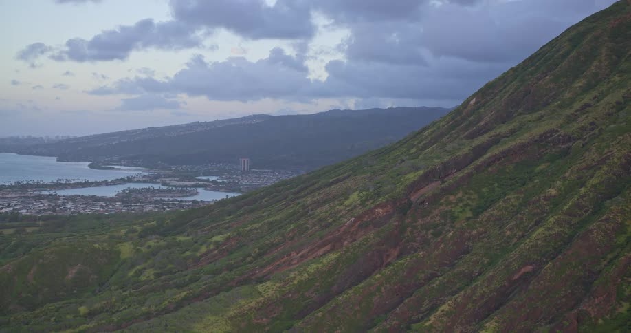 A scenic landscape view of a vast, verdant mountainside overlooking a coastal city under an overcast sky
