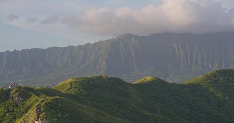 A breathtaking aerial pullback reveals hikers on a lush green mountain ridge in Hawaii, with a majestic mountain range in the distance