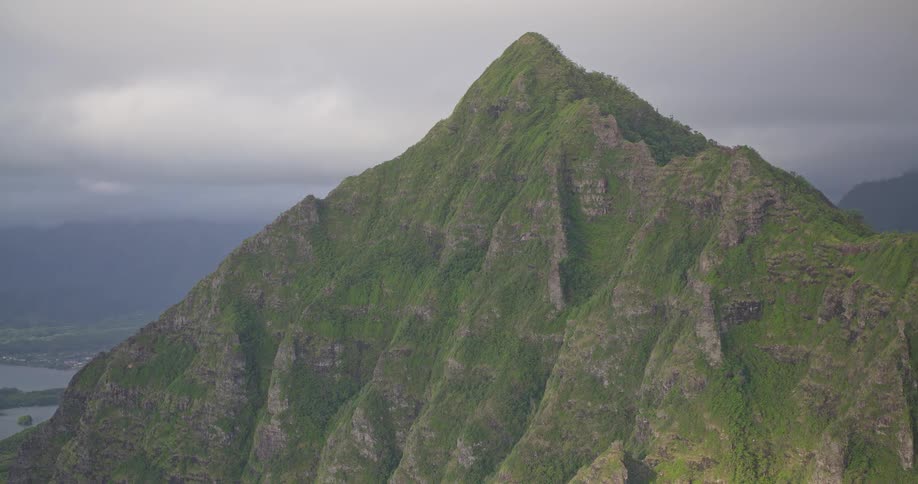 A slow, dramatic zoom on a jagged, lush green mountain peak in Oahu, Hawaii, under a cloudy, overcast sky