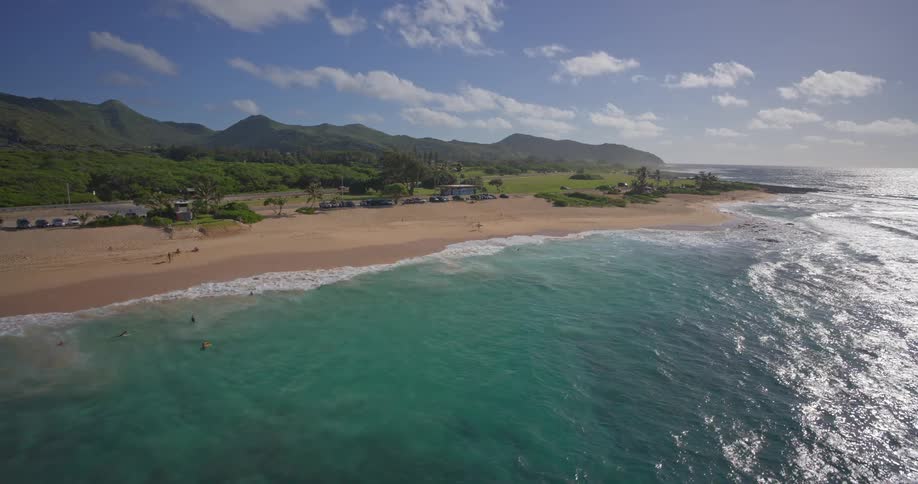 An idyllic aerial shot flies over the beautiful turquoise ocean as waves crash onto a sandy beach with lush green mountains in the background