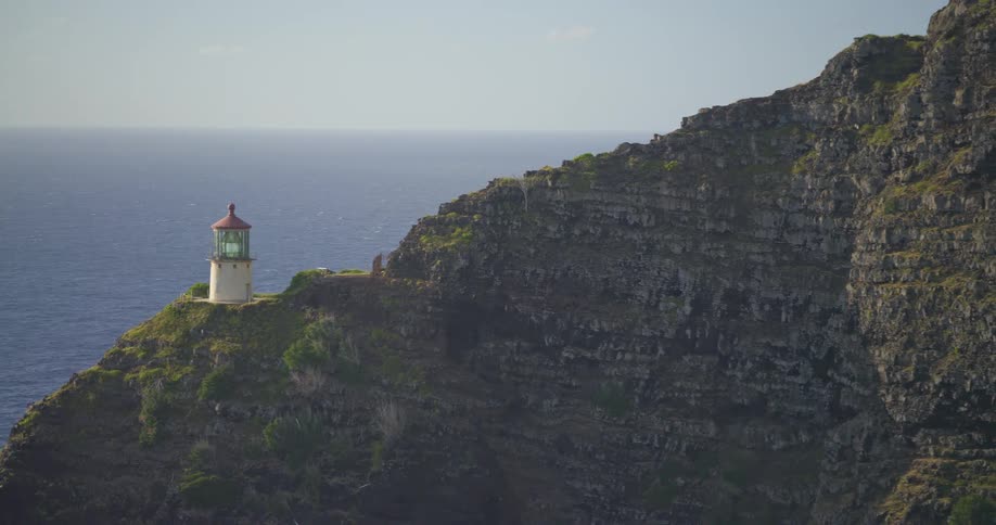 A slow zoom out from a scenic lighthouse perched on a rugged, green cliff overlooking the vast blue ocean