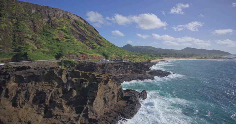 An aerial shot slowly pushes in on a dramatic volcanic coastline on a sunny day, revealing a scenic highway and crashing turquoise ocean waves