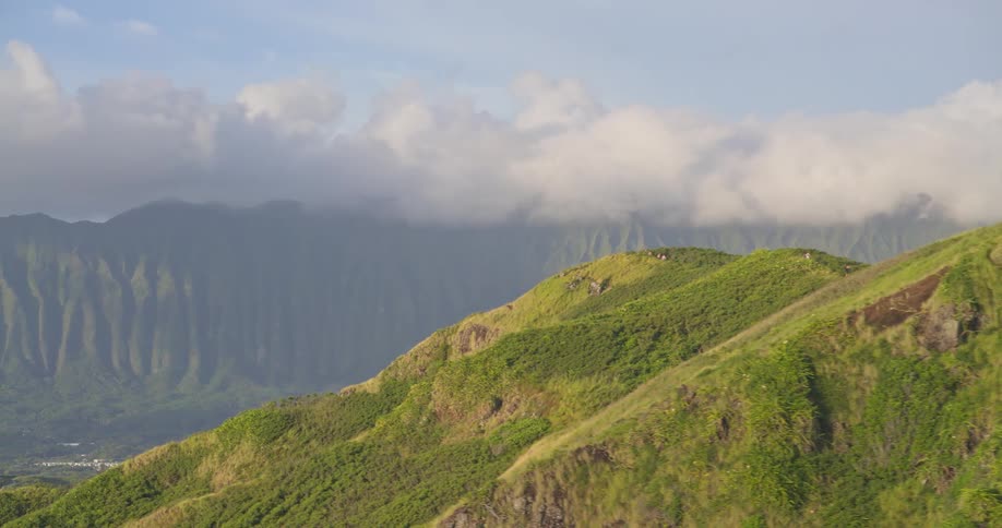 An aerial view pans left along a lush green mountain ridge, revealing hikers at scenic lookout points and a vast mountain range in the background