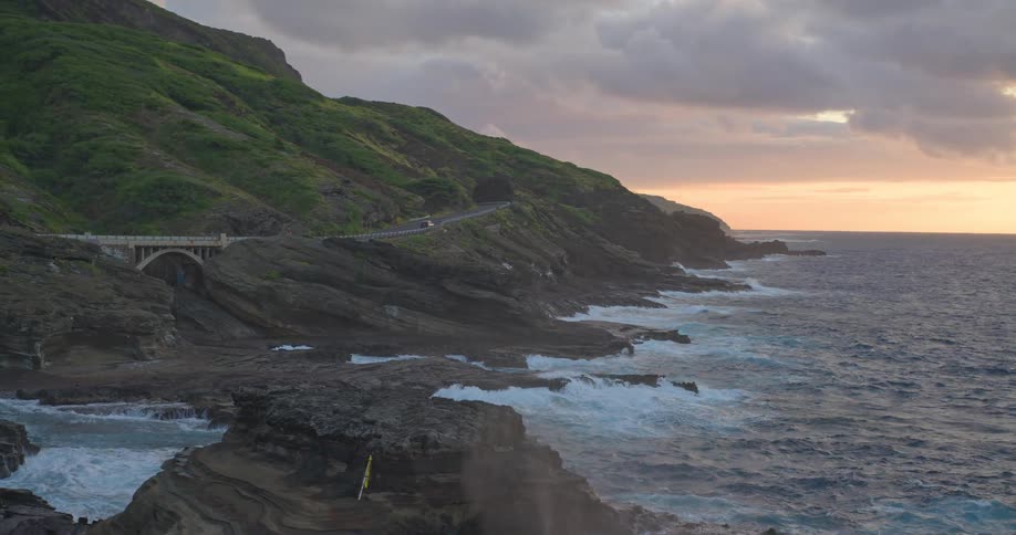 A scenic coastal highway winds along a lush, rocky cliff as powerful ocean waves crash against the shore during a dramatic sunrise