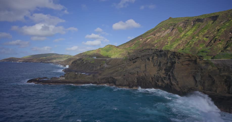A slow pull-back aerial shot reveals the dramatic volcanic coastline of Oahu, Hawaii, as waves crash against the rocks and cars drive on the scenic highway