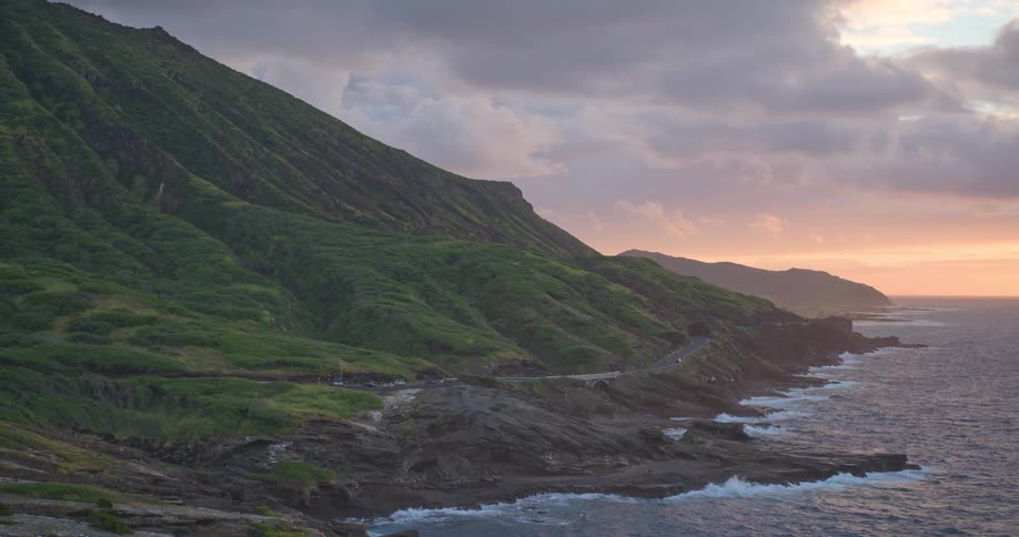 An aerial view captures the dramatic green mountains and rocky coastline of Oahu, Hawaii, as cars drive along the scenic road at sunset