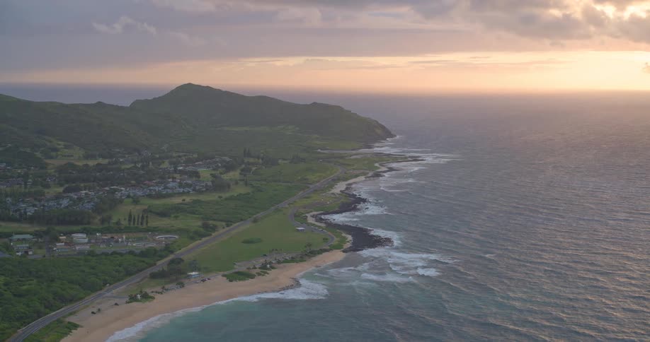 A static aerial view shows a lush tropical island coastline with gentle waves breaking on the shore during a beautiful sunset