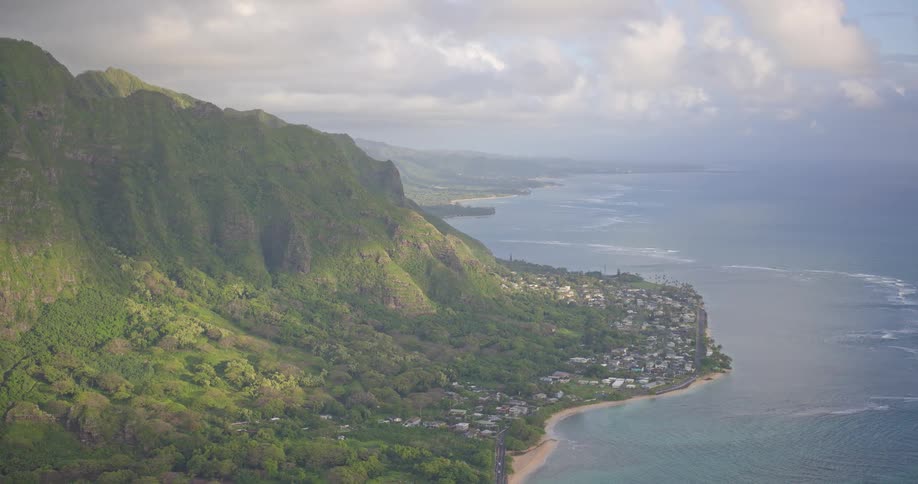 A stunning aerial view pulls back to reveal Hawaii's dramatic green mountains meeting the tranquil Pacific Ocean along a coastal town