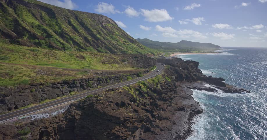 A breathtaking aerial shot captures the stunning beauty of a coastal highway winding between vibrant green cliffs and the sparkling blue ocean