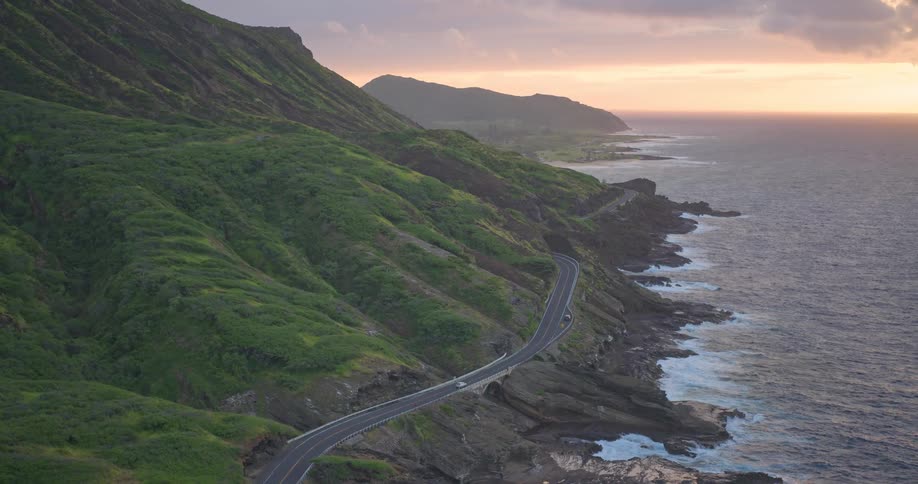 A scenic aerial view shows cars driving along a winding coastal highway nestled between lush green mountains and the ocean at sunrise